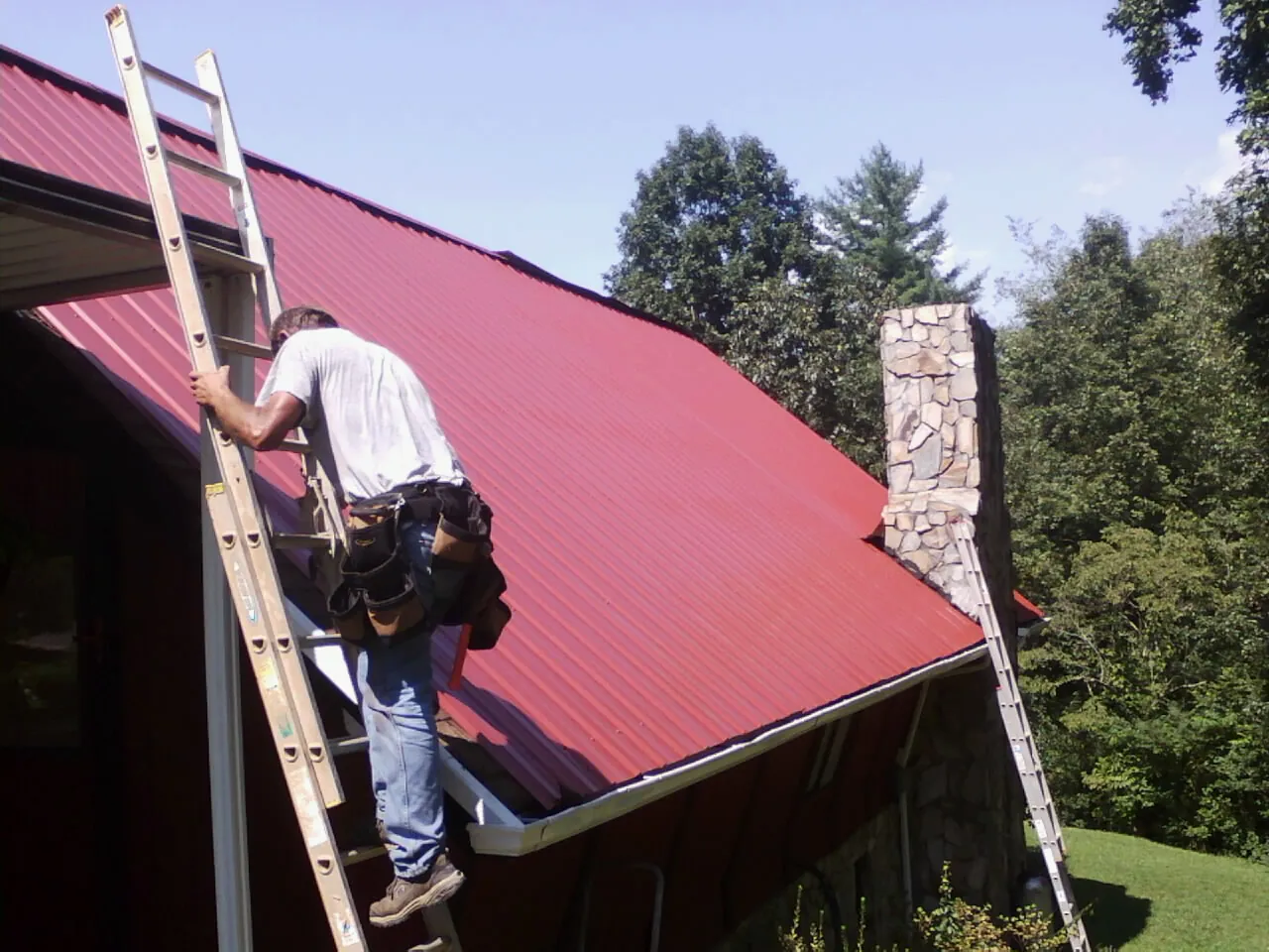 Roofer on a ladder beside a red metal roof and stone chimney — Soffit & Fascia Repair by Roofing Brothers in Salina